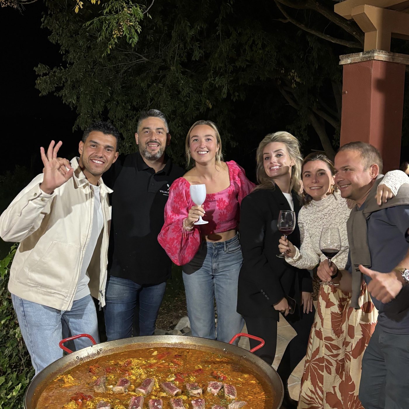 Een groep gasten lacht naar de camera bij een grote pan paella, buiten in de avond